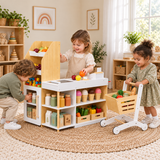 Three children playing shop with a wooden toy grocery store and shopping trolley in a neutral playroom