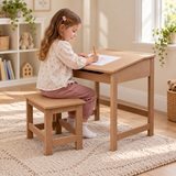 A young girl sits on a small wooden stool at a matching wooden writing desk, drawing on a white sheet of paper in a bright, sunlit room with shelves, plants, and a window in the background.