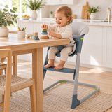 A toddler sits in a high chair at a wooden dining table in a bright, modern kitchen. The high chair has a light gray cushioned seat and blue-gray plastic frame and tray.