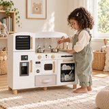 Three-year-old girl stirring a toy pot at a white wooden kitchen in the Little Helper playroom.