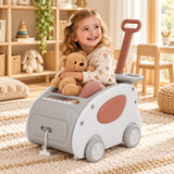 A young girl with blonde hair sits in a toy stroller walker, holding a teddy bear. The stroller walker is white and gray with a dusty rose accent panel and a brown handle.