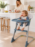 18 month old toddler self feeding in blue/ grey baby high chair with mum in kitchen