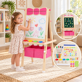 Child drawing on a pink wooden easel with paint pots and storage